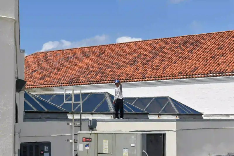 A technician stands on top of a large packaged rooftop unit, checking it over, with blue rooflights and a red barrel-tile roof rising behind him