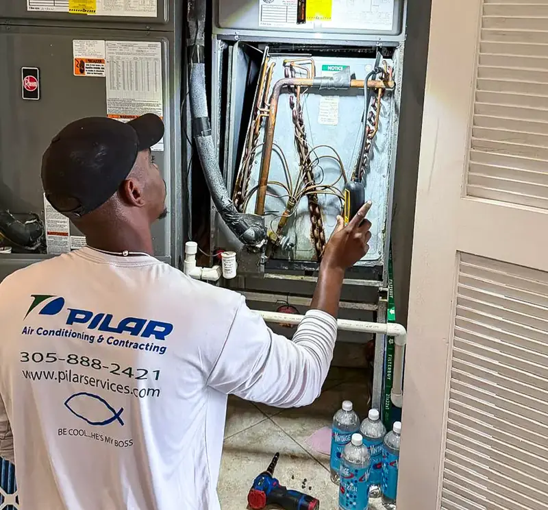 A Pilar Services technician in a branded shirt points up into an open indoor air-handler cabinet, the copper line set, wiring and drain connections exposed and the louvred closet door standing open beside him